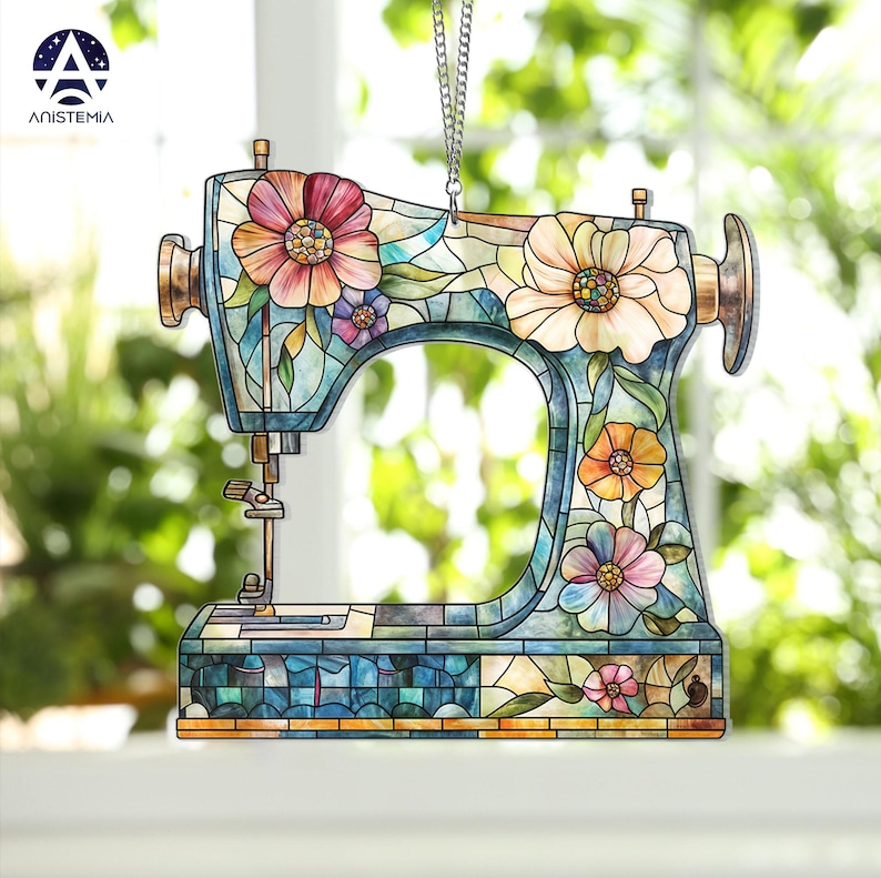 May include: A decorative sewing machine ornament with a stained glass effect. The ornament features a floral design with pink, yellow, and purple flowers. The sewing machine is blue and has a gold-colored handle and needle.