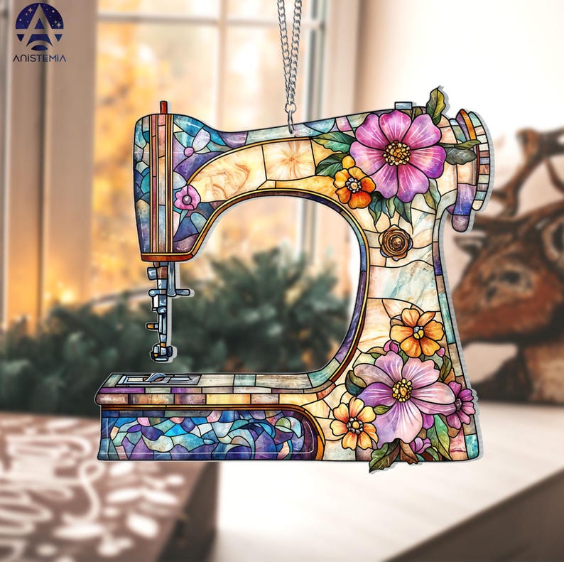 May include: A decorative sewing machine ornament with a stained-glass effect. The ornament features a colorful design with purple, orange, and yellow flowers. The text "ANISTEMIA" is visible in the upper left corner. The ornament hangs from a silver chain.