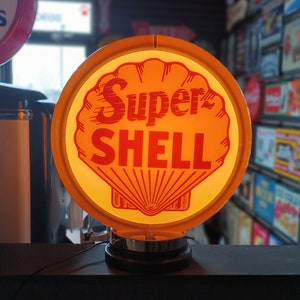 May include: A vintage Super Shell gas station sign with a yellow shell and red lettering. The sign is illuminated and has a black base.