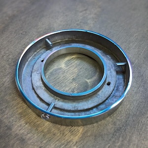 May include: A silver metal ring with a smaller ring in the center. The outer ring has a smooth, polished finish, while the inner ring has a rougher, textured surface. There are three small screws visible on the outer ring.