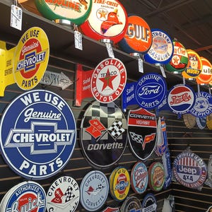 K&ouml;nnte beinhalten: Eine Wandanzeige mit Vintage-Metallschilden mit verschiedenen Marken und Logos, darunter Chevrolet, Texaco, Gulf, Ford, Jeep und Corvette. Die Schilder sind in verschiedenen Farben, darunter Rot, Blau, Gelb und Gr&uuml;n.