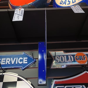 K&ouml;nnte beinhalten: Eine Sammlung von Vintage-Metallschilden mit verschiedenen Marken und Themen, darunter Gulf, Aviation Gasoline, Service, Sold Gulf und Corvette. Die Schilder sind an einer schwarzen Lattenwand mit blauen Metallhalterungen angebracht.