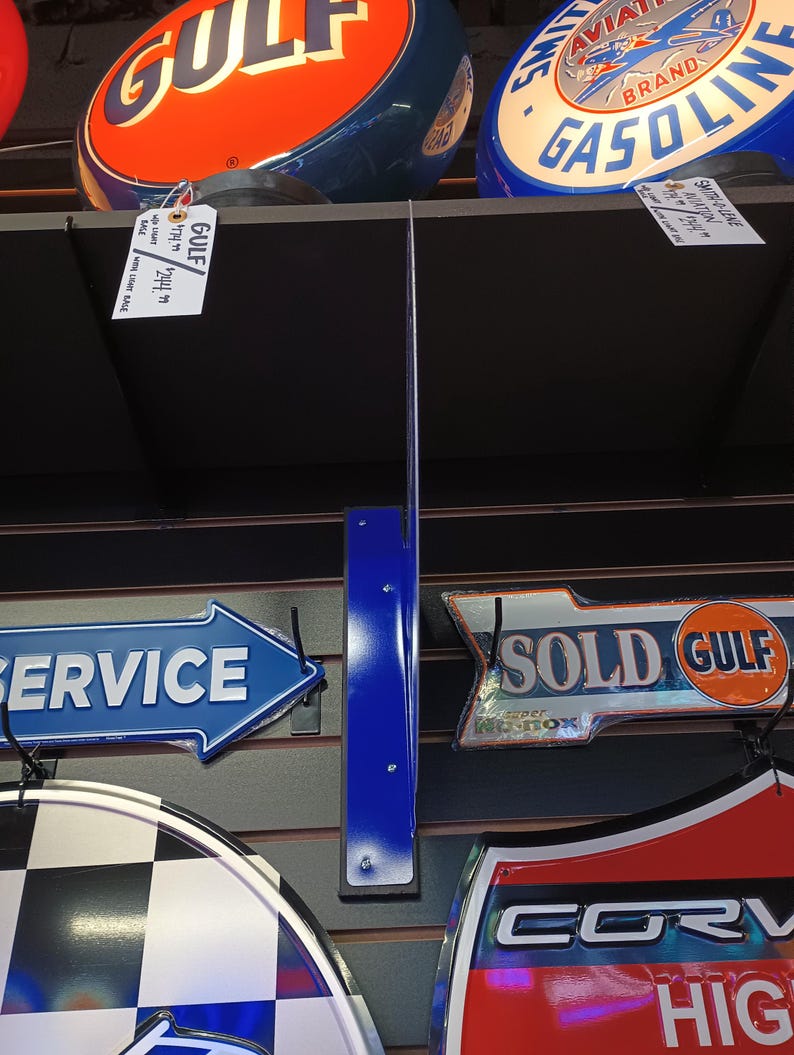 K&ouml;nnte beinhalten: Eine Sammlung von Vintage-Metallschilden mit verschiedenen Marken und Themen, darunter Gulf, Aviation Gasoline, Service, Sold Gulf und Corvette. Die Schilder sind an einer schwarzen Lattenwand mit blauen Metallhalterungen angebracht.