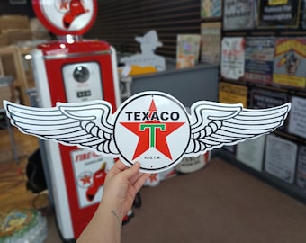 Könnte beinhalten: Ein weißes Metallschild mit einem roten Stern und den Worten "Texaco" und "Reg. T.M." Der Stern hat eine grüne Mitte und ein weißes "T" in der Mitte. Das Schild hat weiße Flügel auf beiden Seiten.