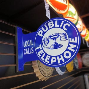 K&ouml;nnte beinhalten: Ein blaues und wei&szlig;es Vintage-Telefon-Schild mit dem Text "PUBLIC TELEPHONE" und "LOCAL CALLS". Das Schild ist an einer Holzwand mit anderen Vintage-Schildern angebracht.
