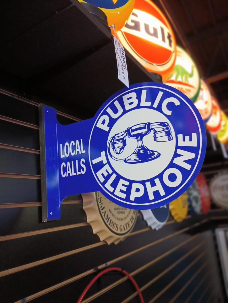 K&ouml;nnte beinhalten: Ein blaues und wei&szlig;es Vintage-Telefon-Schild mit dem Text "PUBLIC TELEPHONE" und "LOCAL CALLS". Das Schild ist an einer Holzwand mit anderen Vintage-Schildern angebracht.