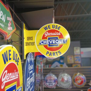 K&ouml;nnte beinhalten: Ein rundes Schild in Gelb und Rot mit dem Text "We Use Genuine Chevrolet Parts". Das Schild h&auml;ngt an einer schwarzen Metallhalterung an einer Wand mit anderen Schildern.
