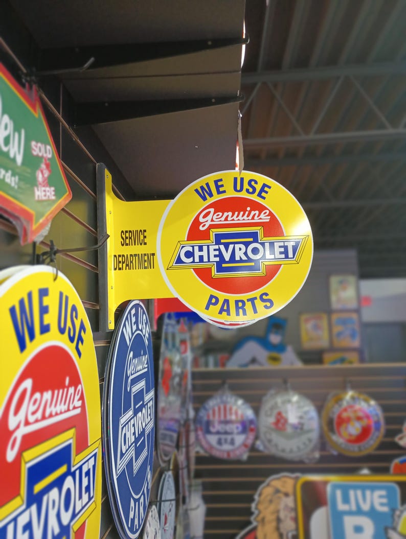 K&ouml;nnte beinhalten: Ein rundes Schild in Gelb und Rot mit dem Text "We Use Genuine Chevrolet Parts". Das Schild h&auml;ngt an einer schwarzen Metallhalterung an einer Wand mit anderen Schildern.