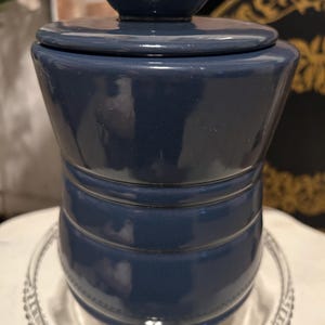 Peut inclure: Un pot en céramique bleu marine avec un couvercle, posé sur une assiette en verre transparent. Le pot a une forme cylindrique avec des rainures horizontales et un petit bouton sur le couvercle. Le pot mesure environ 13 cm de haut.