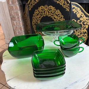 Puede incluir: Una colección de fuentes de vidrio verde vintage. El juego incluye una fuente rectangular, cuencos cuadrados y tazas de té. Los platos están colocados sobre una mesa blanca desgastada, con una chimenea al fondo.