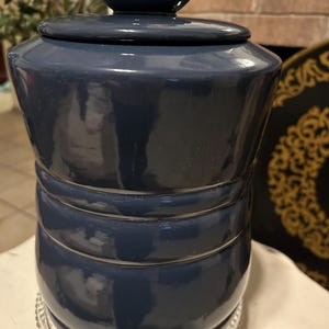 May include: A navy blue ceramic jar with a lid, featuring a glossy finish. The jar has a cylindrical shape with horizontal bands and sits on a clear, decorative glass plate. The lid has a small, rounded handle.