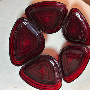 Puede incluir: Un conjunto de cinco platos triangulares de vidrio rojo rubí dispuestos en círculo. Cada plato presenta un diseño geométrico en capas con un interior rojo oscuro y un acabado brillante. Los platos se exhiben sobre una superficie blanca.