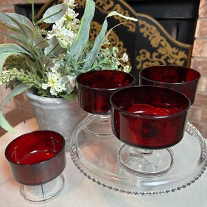 Op de afbeelding: Vier robijnrode glazen dessertschalen op een heldere glazen serveerschaal. Eén schaal is apart. De schalen hebben een afgeronde komvorm en staan op een korte steel. Een potplant staat op de achtergrond.