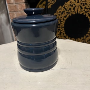 May include: A dark blue ceramic jar with a lid. The jar has a cylindrical shape with horizontal ridges and a small, round lid. The jar is sitting on a white surface. The background includes a brick wall and a decorative black and gold object.