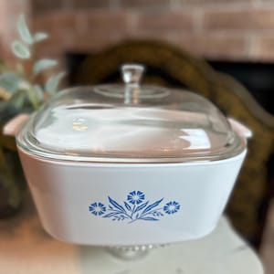 May include: A white casserole dish with a clear glass lid and a clear glass handle. The dish has a blue floral design on the front. The dish is sitting on a clear glass pedestal.