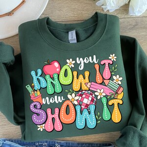 K&ouml;nnte beinhalten: Gr&uuml;nes Sweatshirt mit einem bunten Grafikdesign, auf dem "You Know It Now Show It" steht. Die Grafik enth&auml;lt einen roten Apfel, Bleistifte, ein Lineal, eine Blume und einen karierten Ball.
