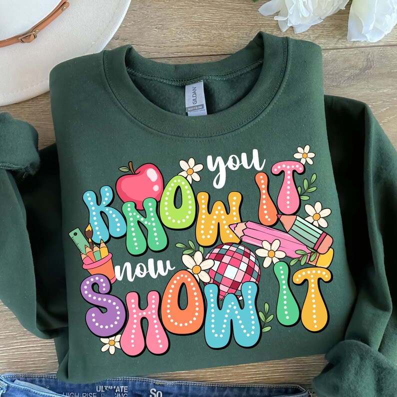 K&ouml;nnte beinhalten: Gr&uuml;nes Sweatshirt mit einem bunten Grafikdesign, auf dem "You Know It Now Show It" steht. Die Grafik enth&auml;lt einen roten Apfel, Bleistifte, ein Lineal, eine Blume und einen karierten Ball.