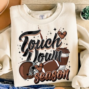 May include: A beige sweatshirt with a brown, black, and white graphic design. The graphic features a football helmet, a football, a coffee cup, and the text "Touch Down Season".