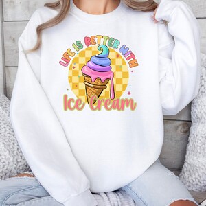 Puede incluir: Sudadera blanca con un gr&aacute;fico de cono de helado colorido y el texto "Life is better with ice cream".