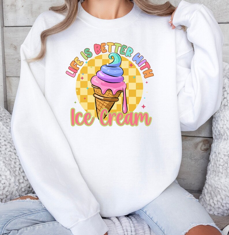 Puede incluir: Sudadera blanca con un gr&aacute;fico de cono de helado colorido y el texto "Life is better with ice cream".