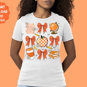 May include: White t-shirt with a design of orange and white bows, a red apple, a globe, a pencil, a coffee cup, and a stack of books. The design is surrounded by small white flowers.