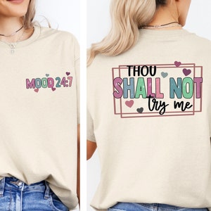 May include: A beige t-shirt with the text "MOOD 24:7" on the front and "THOU SHALL NOT try me" on the back. The text is in a colorful, playful font with hearts.