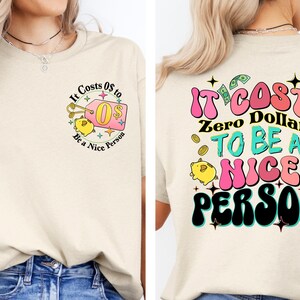May include: A beige t-shirt with a graphic on the front that says "It Costs 0$ to Be a Nice Person" and a piggy bank with a price tag that says "0$". The back of the shirt has a graphic that says "It Costs Zero Dollars to Be a Nice Person" in colorful, retro-style lettering.