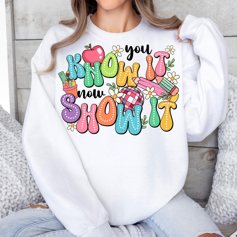 K&ouml;nnte beinhalten: Wei&szlig;er Pullover mit einem bunten Grafikdesign, auf dem "You Know It Now Show It" mit einem roten Apfel, Bleistiften und Blumen steht.