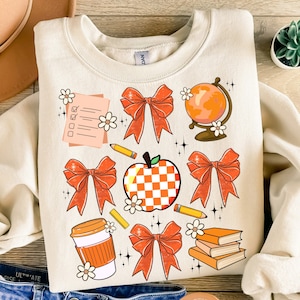 May include: A beige sweatshirt with an orange and white checkered apple, orange bows, a globe, a coffee cup, pencils, books, and a checklist. The design is a cute and whimsical back-to-school theme.