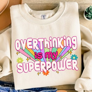 May include: A cream colored sweatshirt with the text "Overthinking is my superpower" in pink and white letters with a rainbow and flowers behind the text.