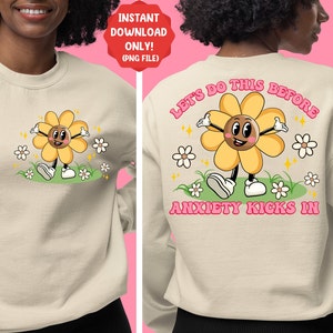 May include: A beige sweatshirt with a cartoon flower graphic on the front and the text "Let's do this before anxiety kicks in" on the back. The flower is yellow with a brown face and is smiling. The text is in pink.