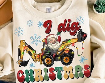 Puede incluir: Una sudadera blanca con una excavadora amarilla con Papá Noel conduciéndola. La excavadora tiene luces de Navidad. El texto "I dig Christmas" está escrito en letras rojas, verdes y amarillas.