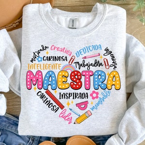 May include: A grey sweatshirt with a colourful graphic design that says "MAESTRA" in a heart shape. The graphic includes words in Spanish describing a teacher, such as "motivada", "creativa", "dedicada", "trabajadora", "inteligente", "cariñosa", "ingeniosa", "inspirada", "memorable", "líder", and "cariñosa".
