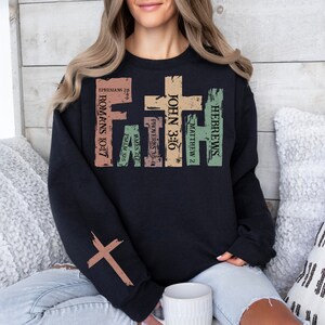 Puede incluir: Sudadera negra con un dise&ntilde;o gr&aacute;fico desgastado que presenta una cruz y vers&iacute;culos b&iacute;blicos, incluyendo "Romanos 10:17", "Juan 3:16", "Hebreos", "Mateo 2", "Proverbios 3:5-6", "Santiago 2:17" y "Efesios 2:8".