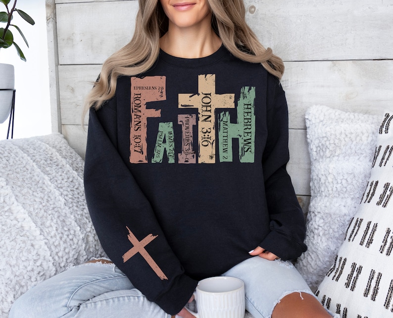 Puede incluir: Sudadera negra con un dise&ntilde;o gr&aacute;fico desgastado que presenta una cruz y vers&iacute;culos b&iacute;blicos, incluyendo "Romanos 10:17", "Juan 3:16", "Hebreos", "Mateo 2", "Proverbios 3:5-6", "Santiago 2:17" y "Efesios 2:8".