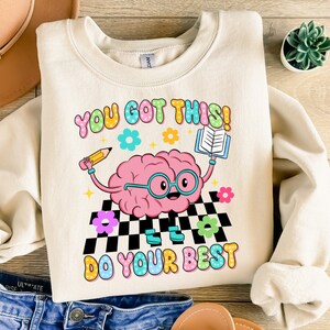 May include: A cream colored sweatshirt with a cartoon brain wearing glasses and holding a pencil and a book. The text "You got this! Do your best" is printed on the sweatshirt in colorful letters.