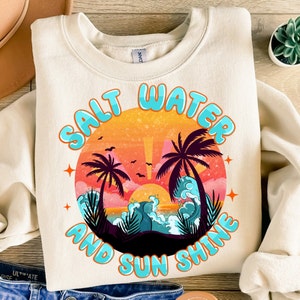May include: A cream colored sweatshirt with a graphic design of a sunset with palm trees and waves. The text "Salt Water and Sun Shine" is in a blue and orange font.