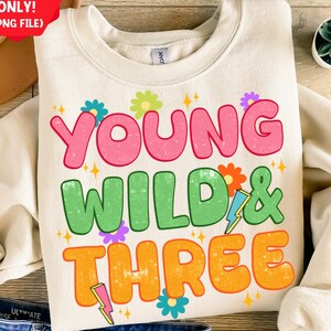 Może przedstawiać: Beżowa bluza z napisem "Young Wild & Three" w kolorowych, retro literach. Tekst jest otoczony małymi kwiatami i błyskawicami.