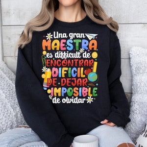 Op de afbeelding: Zwart sweatshirt met een kleurrijk grafisch ontwerp met de tekst "Una gran maestra es difficult de encontrar, dificil de dejar, imposible de olvidar." De grafiek bevat een papieren vliegtuig, een potlood, een bloem, een wereldbol, een appel en een smiley.