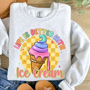 Puede incluir: Una sudadera gris con el texto "Life is better with ice cream" en colores arco&iacute;ris. Un cono de helado de dibujos animados con chispas de arco&iacute;ris y helado rosa, morado y azul se encuentra en el centro del texto.