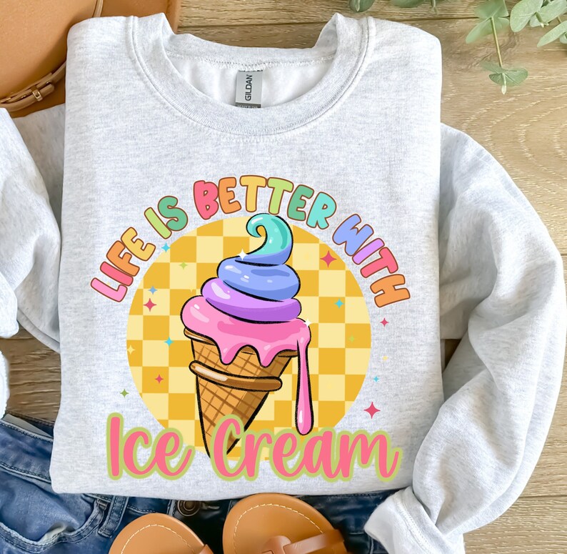 Puede incluir: Una sudadera gris con el texto "Life is better with ice cream" en colores arco&iacute;ris. Un cono de helado de dibujos animados con chispas de arco&iacute;ris y helado rosa, morado y azul se encuentra en el centro del texto.