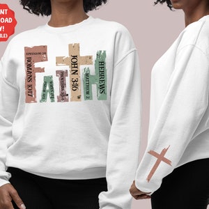 Puede incluir: Sudadera blanca con un dise&ntilde;o gr&aacute;fico desgastado de la palabra "FAITH" con vers&iacute;culos b&iacute;blicos. El gr&aacute;fico est&aacute; en tonos de marr&oacute;n, verde y beige. La sudadera tiene un gr&aacute;fico de cruz en la manga.