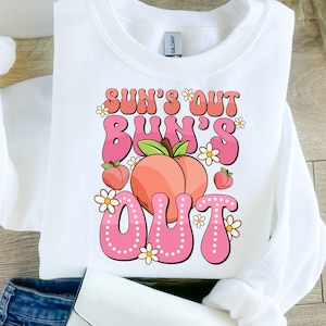 May include: White sweatshirt with pink text that reads "Sun's Out Bun's Out" with a peach graphic and white flowers.