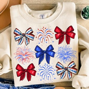 May include: A cream-coloured sweatshirt with a festive design featuring red, white and blue bows and fireworks. The bows are tied with a ribbon and the fireworks are exploding in the air.