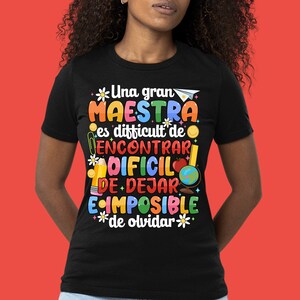 Op de afbeelding: Zwart T-shirt met een kleurrijk grafisch ontwerp met de tekst "Una gran MAESTRA es difficult de ENCONTRAR DIFCIL DE DEJAR E IMPOSIBLE de olvidar" met illustraties van schoolbenodigdheden, een wereldbol en bloemen.