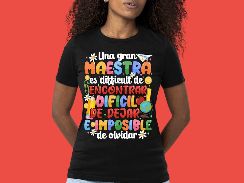 Op de afbeelding: Zwart T-shirt met een kleurrijk grafisch ontwerp met de tekst "Una gran MAESTRA es difficult de ENCONTRAR DIFCIL DE DEJAR E IMPOSIBLE de olvidar" met illustraties van schoolbenodigdheden, een wereldbol en bloemen.