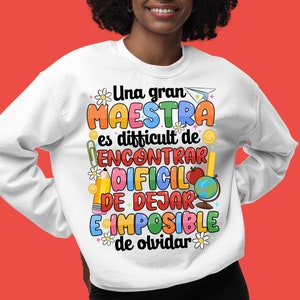 Op de afbeelding: Wit sweatshirt met een kleurrijk grafisch ontwerp met de tekst "Una gran maestra es difficult de encontrar, dificil de dejar, e imposible de olvidar". De grafiek bevat illustraties van een potlood, een aardbol, een boek en een bloem.