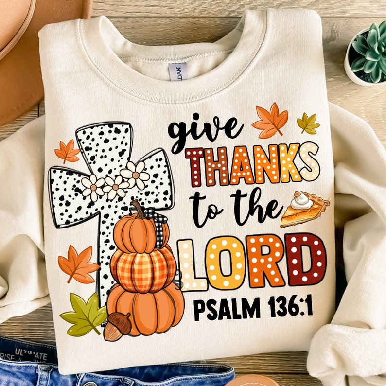 May include: A beige sweatshirt with a graphic design featuring a cross with black and white polka dots, a stack of pumpkins, fall leaves, and the text "give THANKS to the LORD PSALM 136:1".