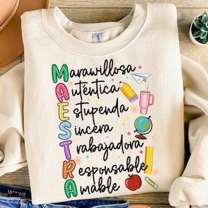 Può includere: Una felpa beige con la parola "MAESTRA" scritta verticalmente in colori arcobaleno. Ogni lettera è decorata con un'immagine diversa a tema scolastico, come una matita, un mappamondo, un righello e una mela. Il testo intorno alle lettere dice "maravillosa, auténtica, estupenda, sincera, trabajadora, responsable, amable".