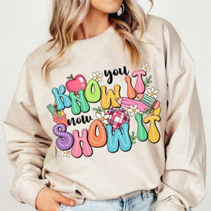 K&ouml;nnte beinhalten: Ein beiger Pullover mit einem bunten Grafikdesign, auf dem "You Know It Now Show It" mit einem roten Apfel, Bleistiften und Blumen steht.